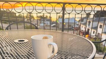 A white mug on a round metal table with a metal railing and a view of a sunset.