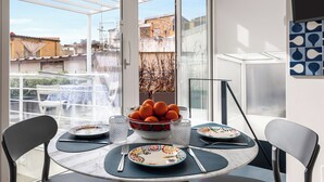 Un coin repas avec une table ronde dressée pour quatre personnes, un saladier rempli d’oranges et une vue sur le paysage urbain à travers de grandes fenêtres.