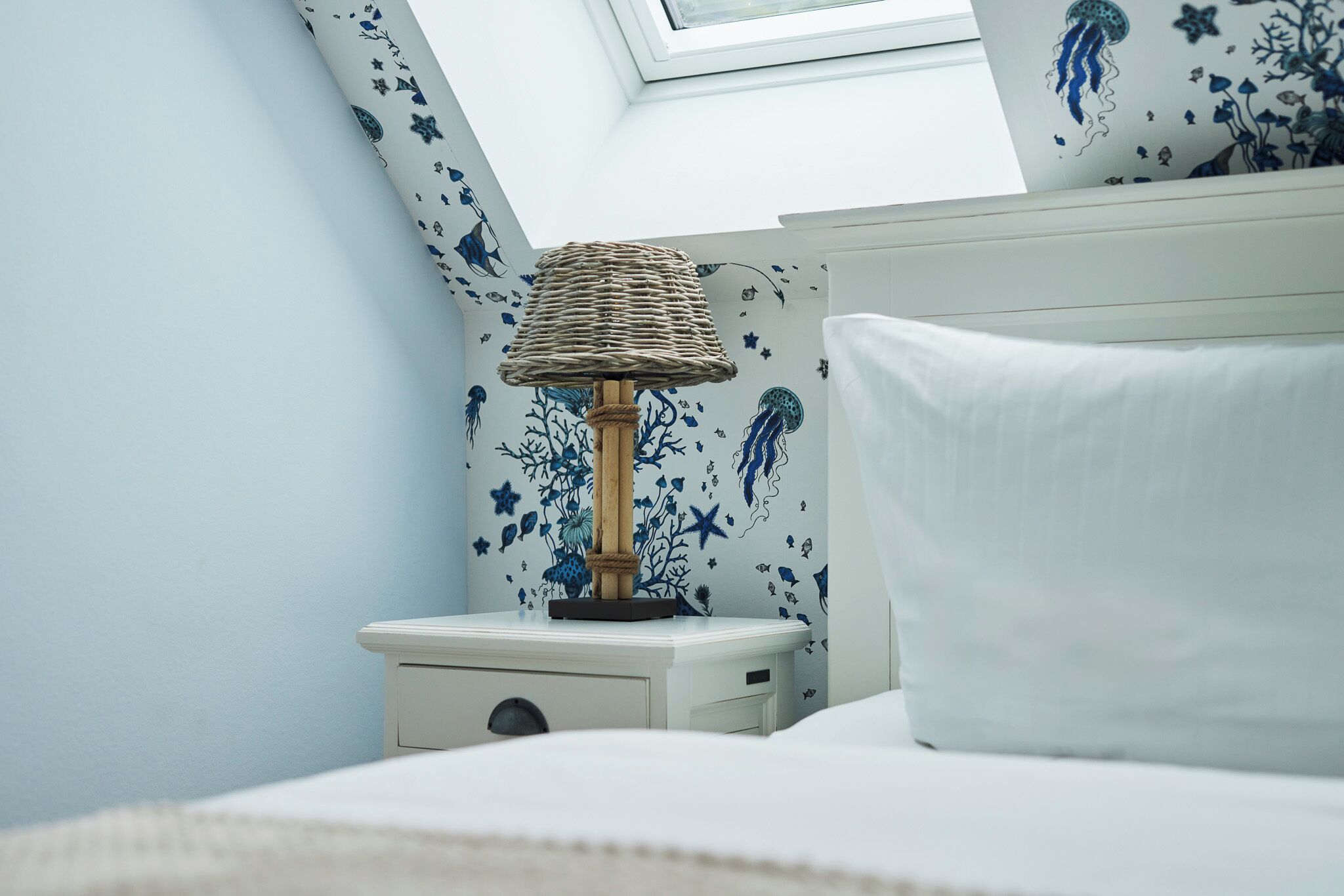A neatly made bed with a white pillow and a bedside table with a lamp. The wall features blue floral wallpaper.