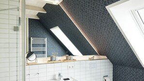 Une salle de bain moderne avec un lavabo blanc, un miroir et un porte-serviettes. Un mur à motifs et un plafond en pente complètent l’ensemble.