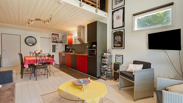 Eine moderne Küche mit roten Schränken, ein Essbereich mit einem roten Tisch, ein Wohnzimmer mit einer Couch und Fernseher sowie ein Balkon mit Holzboden.