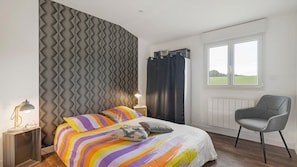 A bedroom with a bed, a chair, a bedside table, a lamp, a window, and a radiator. - Gîte "Le Relais De Jehan" with Private Pool, Terrace, and Garden (Saint-Malo des Trois Fontaines)