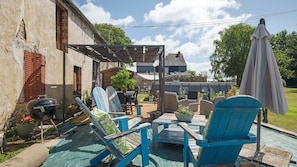 A patio with blue Adirondack chairs, a barbecue grill, and a pergola with a table and chairs underneath. - Gîte "Le Relais De Jehan" with Private Pool, Terrace, and Garden (Saint-Malo des Trois Fontaines)