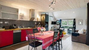 Eine moderne Küche mit Kochinsel, ein Essbereich mit rotem Tischlein und ein Wohnzimmer mit Sofa und Fernseher.