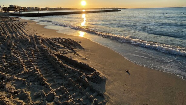 En strand med fodaftryk, der fører ned til vandet, en mole i baggrunden og solnedgang over horisonten.