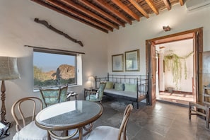 A room with a round table, chairs, a sofa, a bed, a window with a view, and wooden beams on the ceiling.