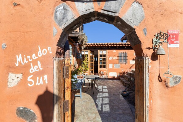 An archway leading to a courtyard with a table and chairs, a bell, and a sign indicating a 24-hour service.