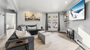 A modern living room with a grey sofa, a coffee table, and a TV. There is a large window with curtains, a door leading to another room, and a fireplace. - Treehouse C106 - Renovated Condo, Pool, Hot Tub, Game Room + Central Summit County Location (Silverthorne)
