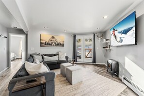 Un salon moderne avec un canapé gris, une table basse et un téléviseur. Il y a une grande fenêtre avec des rideaux, une porte donnant sur une autre pièce et une cheminée.