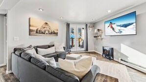 A modern living room with a grey sectional sofa, a fireplace, and two large wall-mounted TVs displaying scenic images. - Treehouse C106 - Renovated Condo, Pool, Hot Tub, Game Room + Central Summit County Location (Silverthorne)