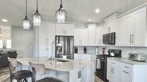 A modern kitchen with white cabinets, stainless steel appliances, and a marble island with bar stools. - Twilight Mist - 3/2 in The Village of Poinciana (Poinciana, The Villages)