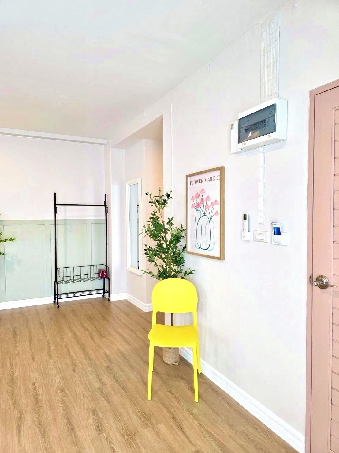 Una habitación con piso de madera, una silla amarilla, un perchero de metal negro, una planta en maceta, un cuadro enmarcado y una puerta con interruptor de luz.