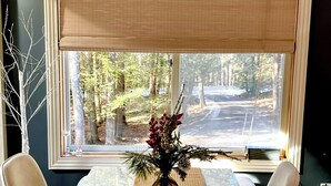 A dining area with a table set for four, a vase with flowers, and a view of a forest through a large window with blinds.