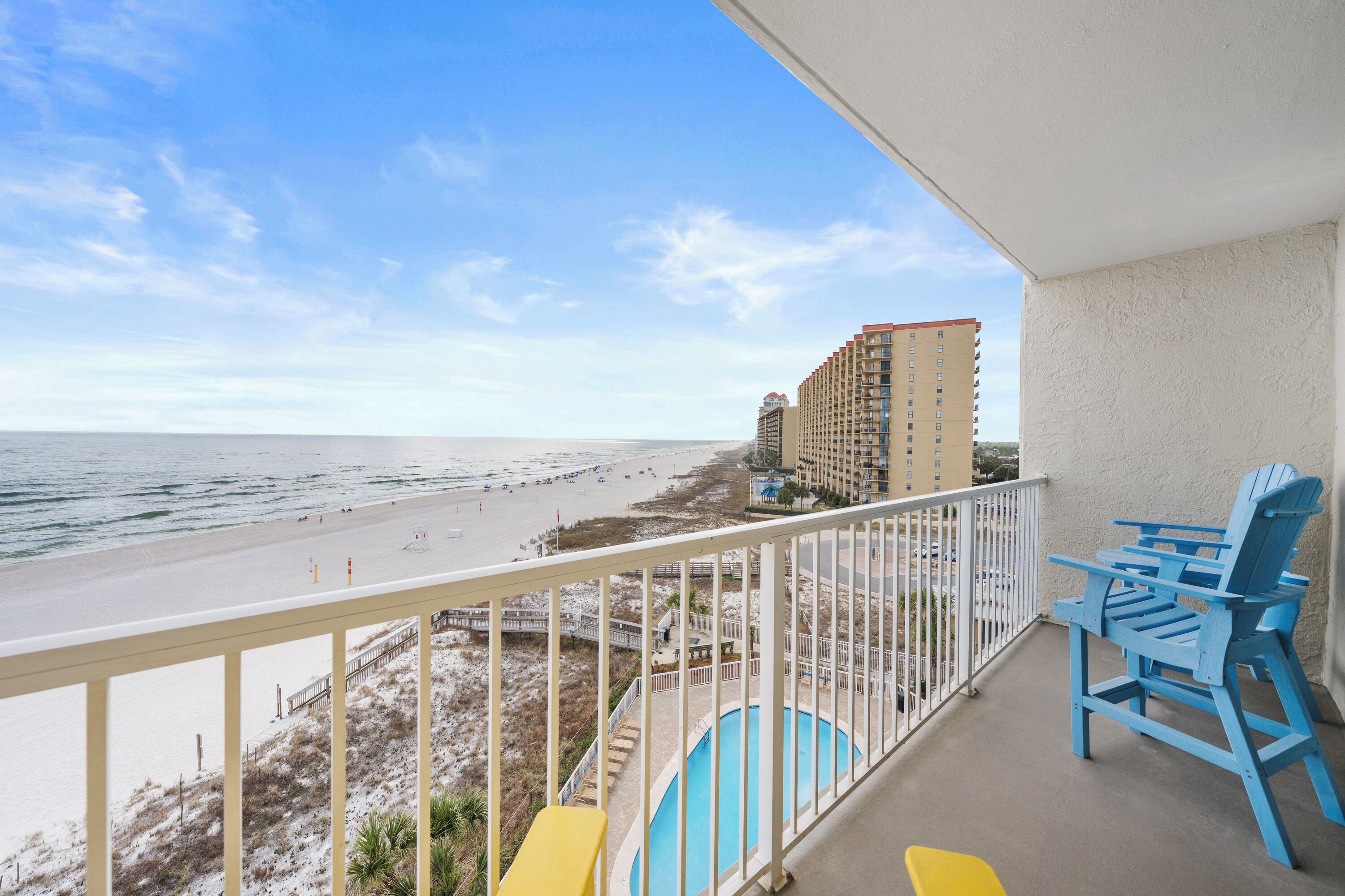 A balcony with a view of the beach and ocean, a pool, and blue and yellow chairs.
