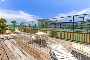 Eine Holzterrasse mit Sitzgelegenheiten im Freien, einem Tennisplatz und Blick auf ein Hotelgebäude.