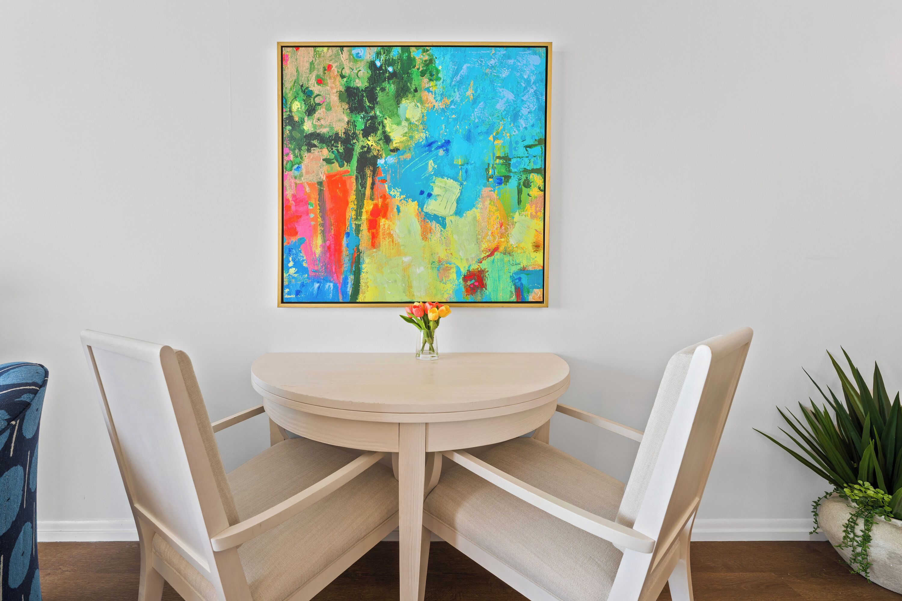 A dining area with a round table, two chairs, a vase with flowers, and a colorful abstract painting on the wall.
