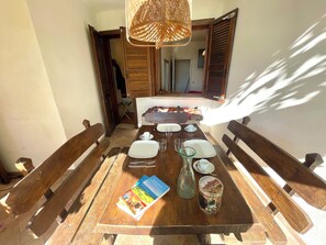 Ein Essbereich mit einem Holztisch, gedeckt für vier Personen, einschließlich Teller, Gläser und Besteck. Eine Korbtreleuchte hängt darüber, und auf dem Tisch liegen ein Buch und eine dekorative Dose.