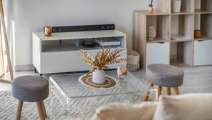 Un salon moderne avec une table basse en verre, deux poufs gris, un téléviseur blanc posé sur une console et une bibliothèque en bois.