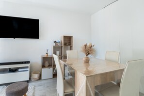 Een moderne woonkamer met een flatscreen televisie aan de muur, een houten eettafel met stoelen, een witte bank en een klein bijzettafeltje met een vaas.