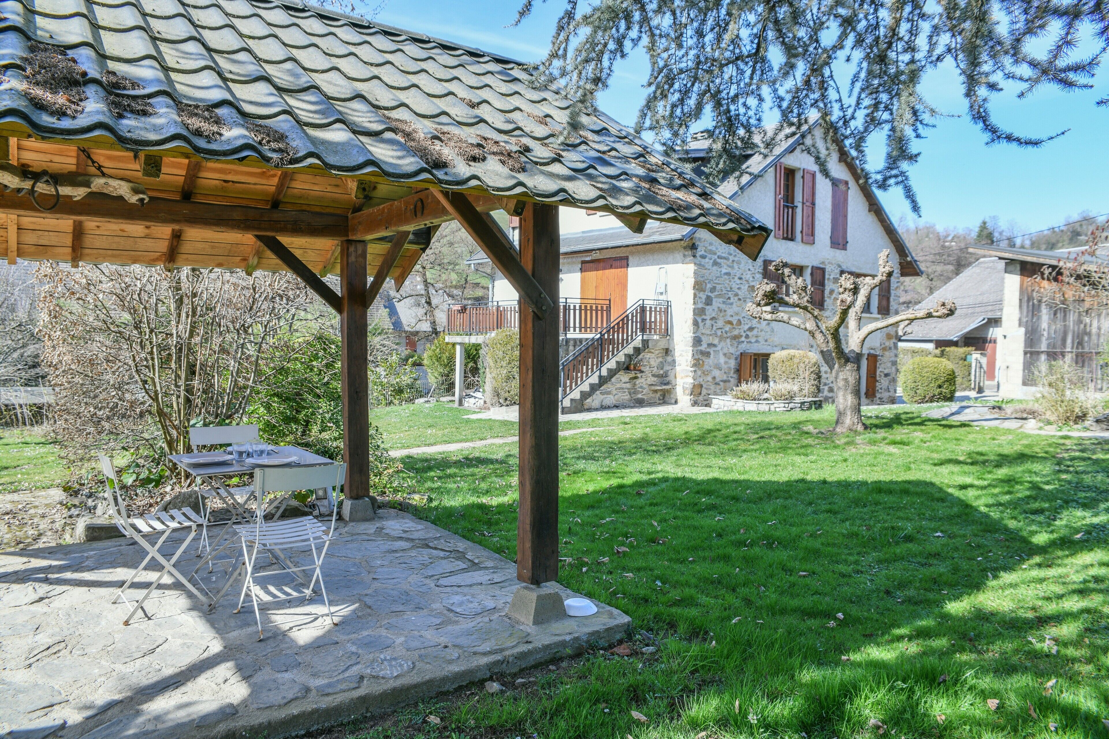 Un patio de piedra con mobiliario exterior, una pérgola de madera y una casa de piedra con techo rojo al fondo.
