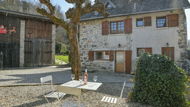 Un bâtiment en pierre avec un arbre devant, une table avec des chaises et une bouteille.