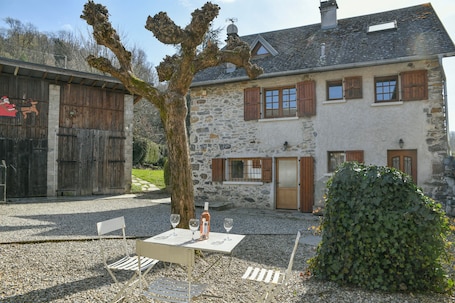 A stone building with a tree in front, a table with chairs, and a bottle.