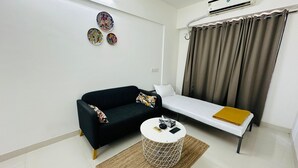 A modern, minimalist living room with a black sofa, a white side table, and a single bed with a brown blanket.