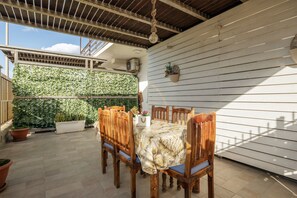 Un patio con una mesa y sillas de madera, rodeado de plantas en macetas y una pared blanca.