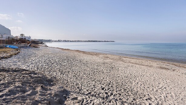 Une plage de sable, un ciel dégagé, une côte lointaine et des bâtiments sur la gauche.