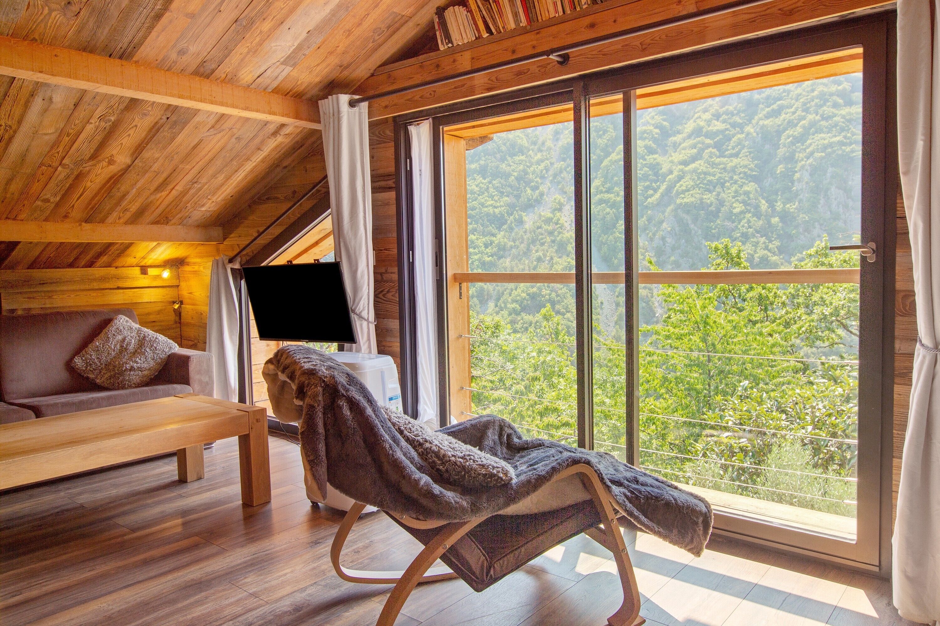 Ein gemütliches Zimmer mit Holzbalkendecke, einer Couch mit Kissen, einem Sessel mit Decke, einem Flachbildfernseher und einem großen Fenster mit Blick auf Grünflächen.