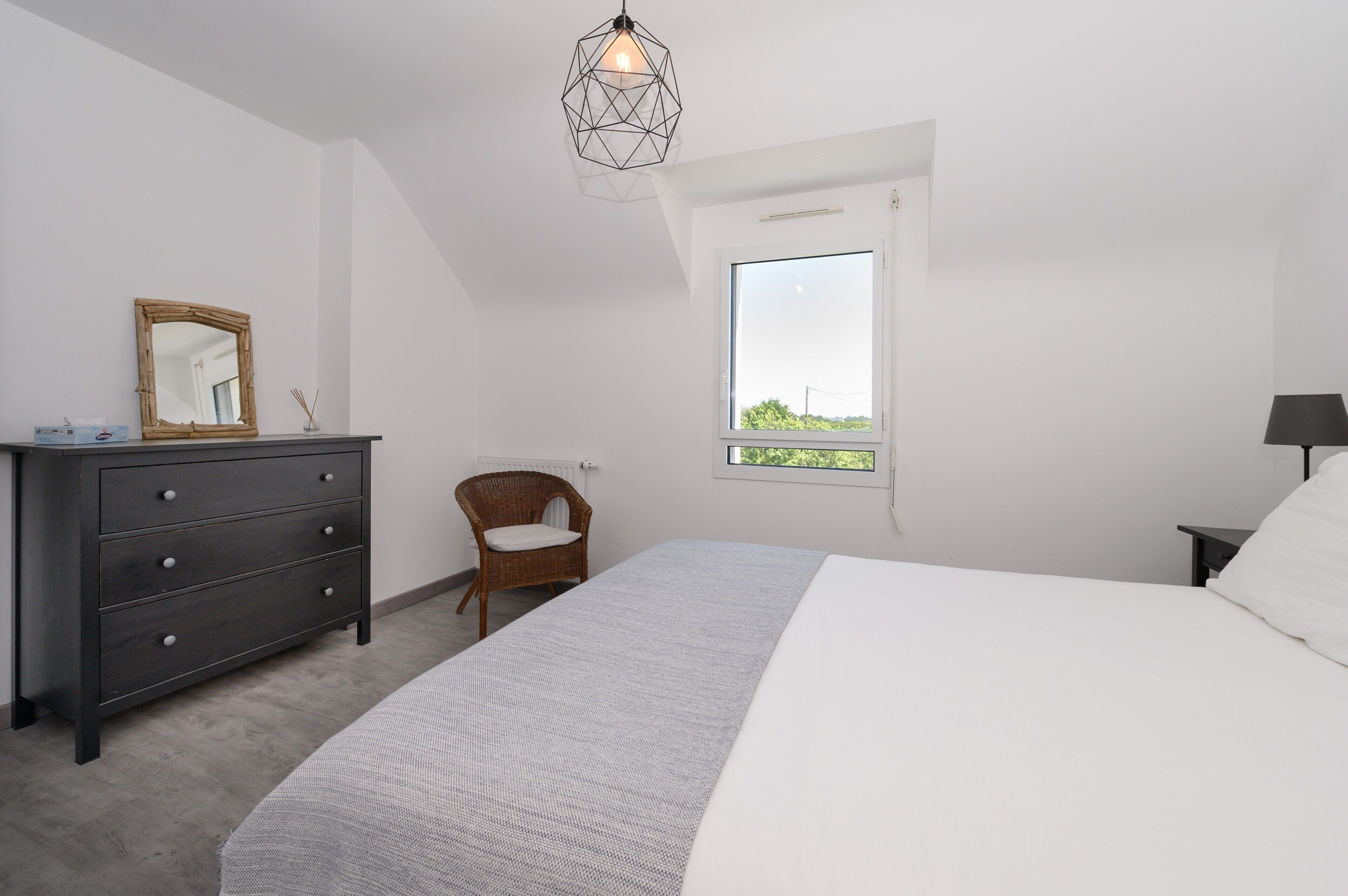 A bedroom with a bed, dresser, chair, window, and geometric pendant light.