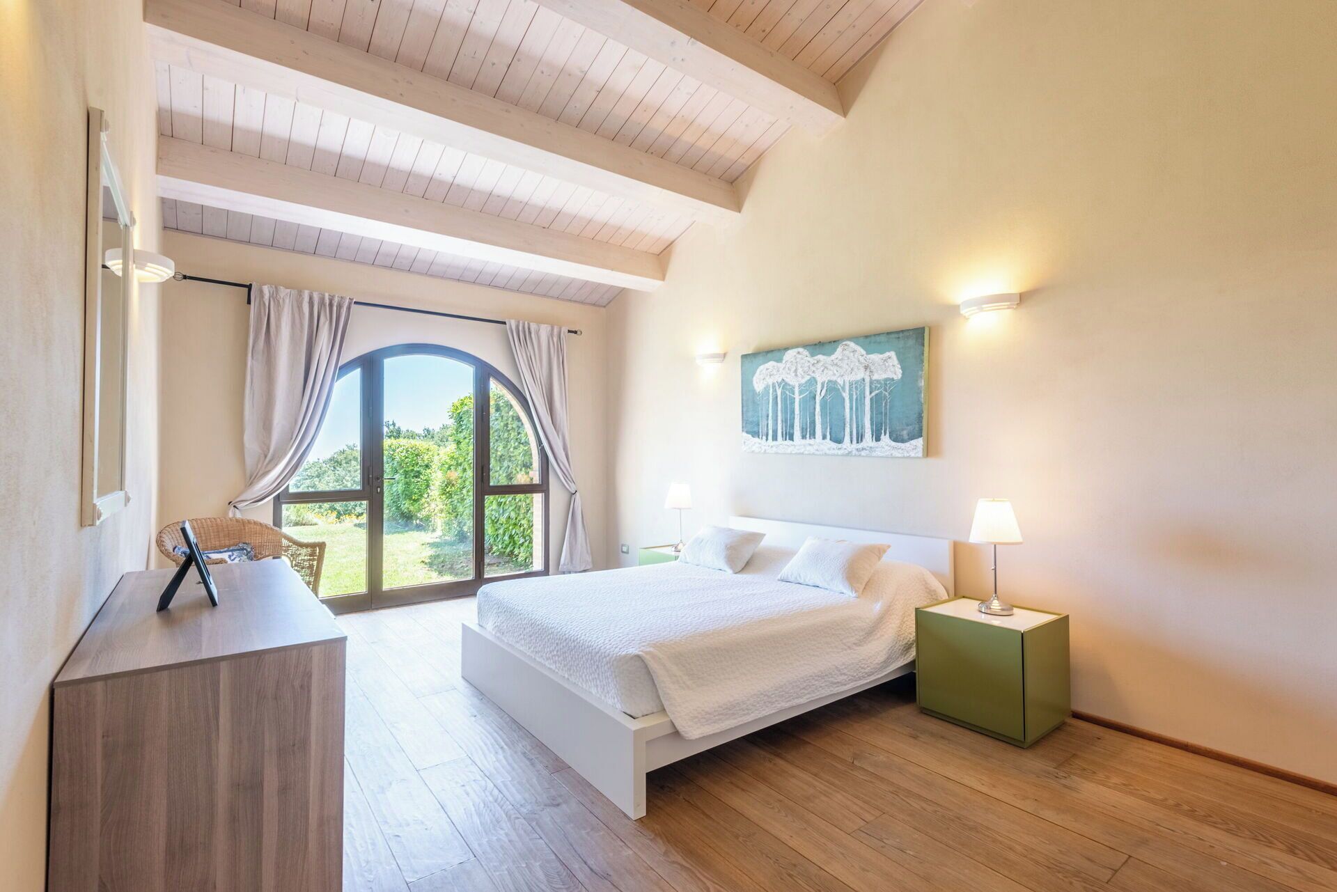 Une chambre à coucher avec un grand lit, un bureau, une chaise, une fenêtre avec des rideaux, un tableau au mur et une table de chevet avec une lampe.
