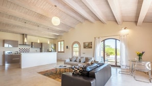 A spacious living area with a sofa, a coffee table, and a dining table. There is a kitchen area with a modern design, including a microwave and a coffee machine. The room has a high ceiling with wooden beams and large windows that provide natural light. - Cosy apartment with WIFI, pool, A/C, TV, patio, pets allowed, panoramic view, close to Montepulciano (Torrita Di Siena)