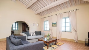 A living room with a sofa, armchairs, a coffee table, and a rug. There are windows with curtains, a door, and a view of the kitchen. - Nice apartment with pool, A/C, WIFI, TV, patio, pets allowed, panoramic view, close to Montepulciano (Torrita Di Siena)