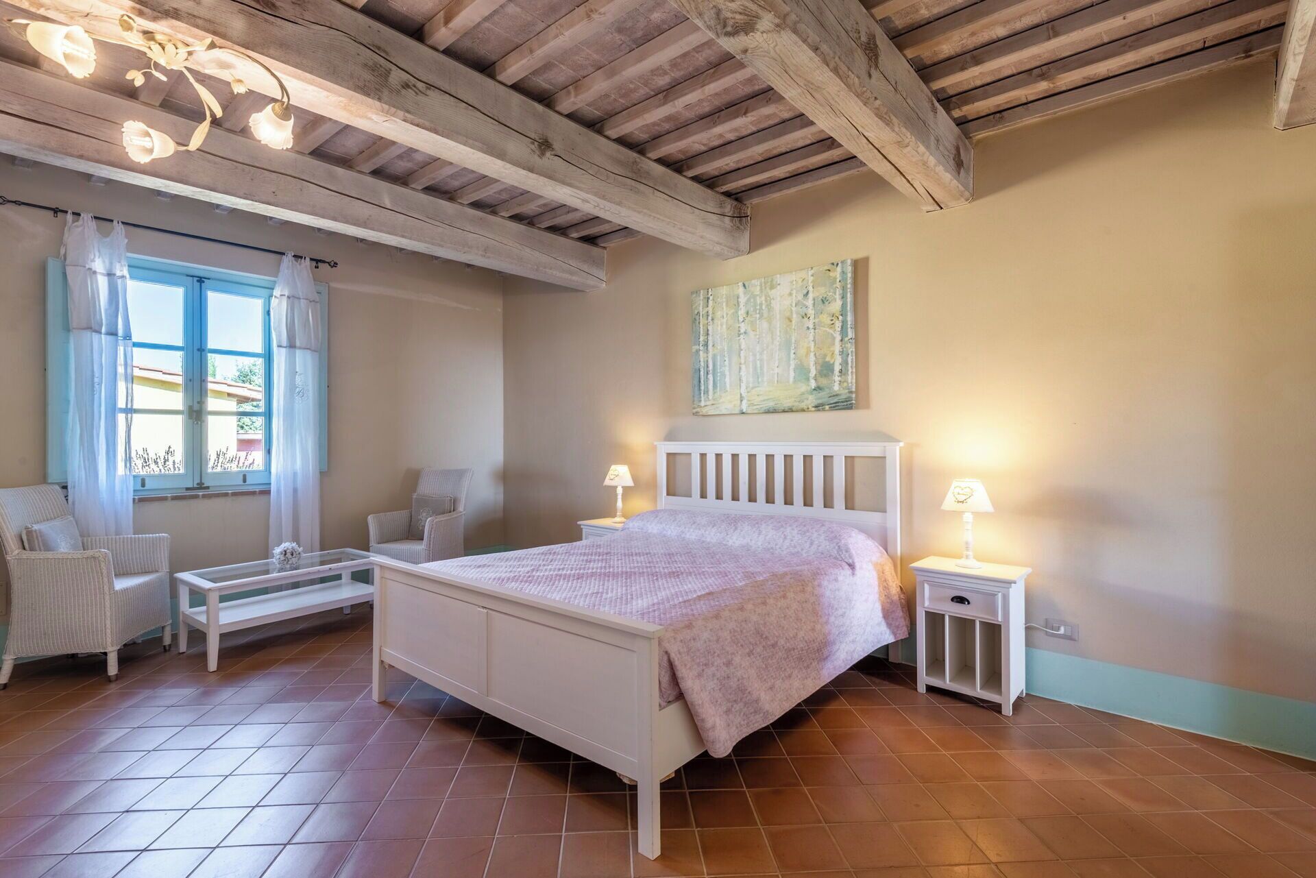 Een slaapkamer met een wit bed, een stoel, een nachtkastje en een raam met gordijnen.