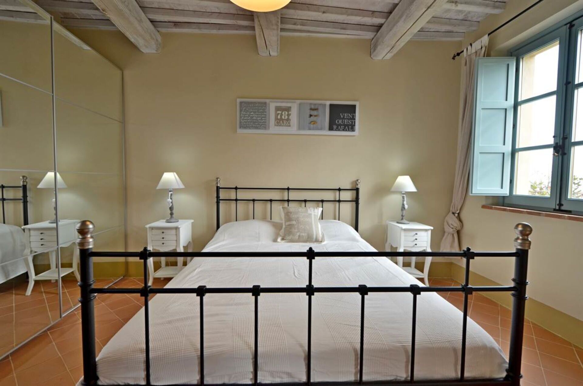 Een slaapkamer met een bed, twee nachtkastjes met lampen, een raam met gordijnen en een spiegel aan de muur.