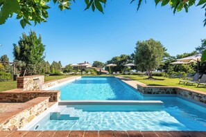 Una piscina rectangular con escalones de piedra para entrar, rodeada de césped, árboles y sombrillas.
