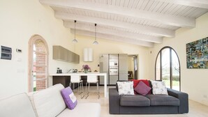 Un saló modern amb un sofà blanc, un sofà gris i una taula de centre. Hi ha una zona de cuina amb taulell blanc i un frigorífic. L'espai té un sostre alt amb bigues a la vista i grans finestres.