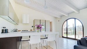 Una cuina moderna amb taulell blanc, tamborets i vista a l'exterior a través d'una gran finestra apuntada.