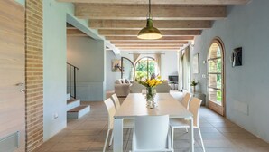 Un comedor amplio con una larga mesa y sillas blancas, una lámpara colgante y vistas a un jardín a través de ventanales arqueados.
