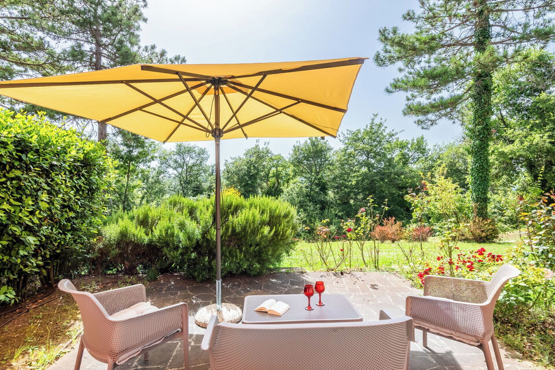 An outdoor seating area with a table and chairs, a yellow umbrella, and a garden with various plants and flowers.