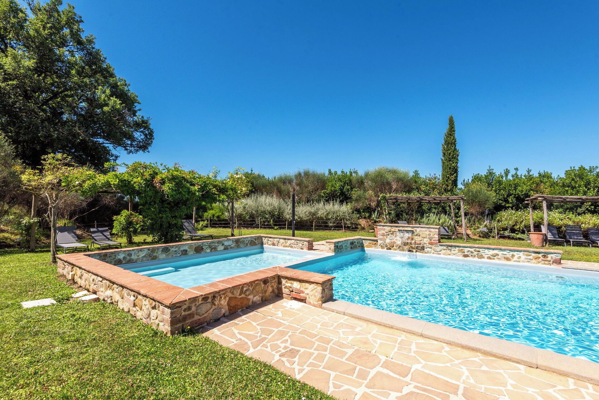 Une piscine rectangulaire bordée de pierres et de briques, entourée d’une pelouse verdoyante et d’arbres.