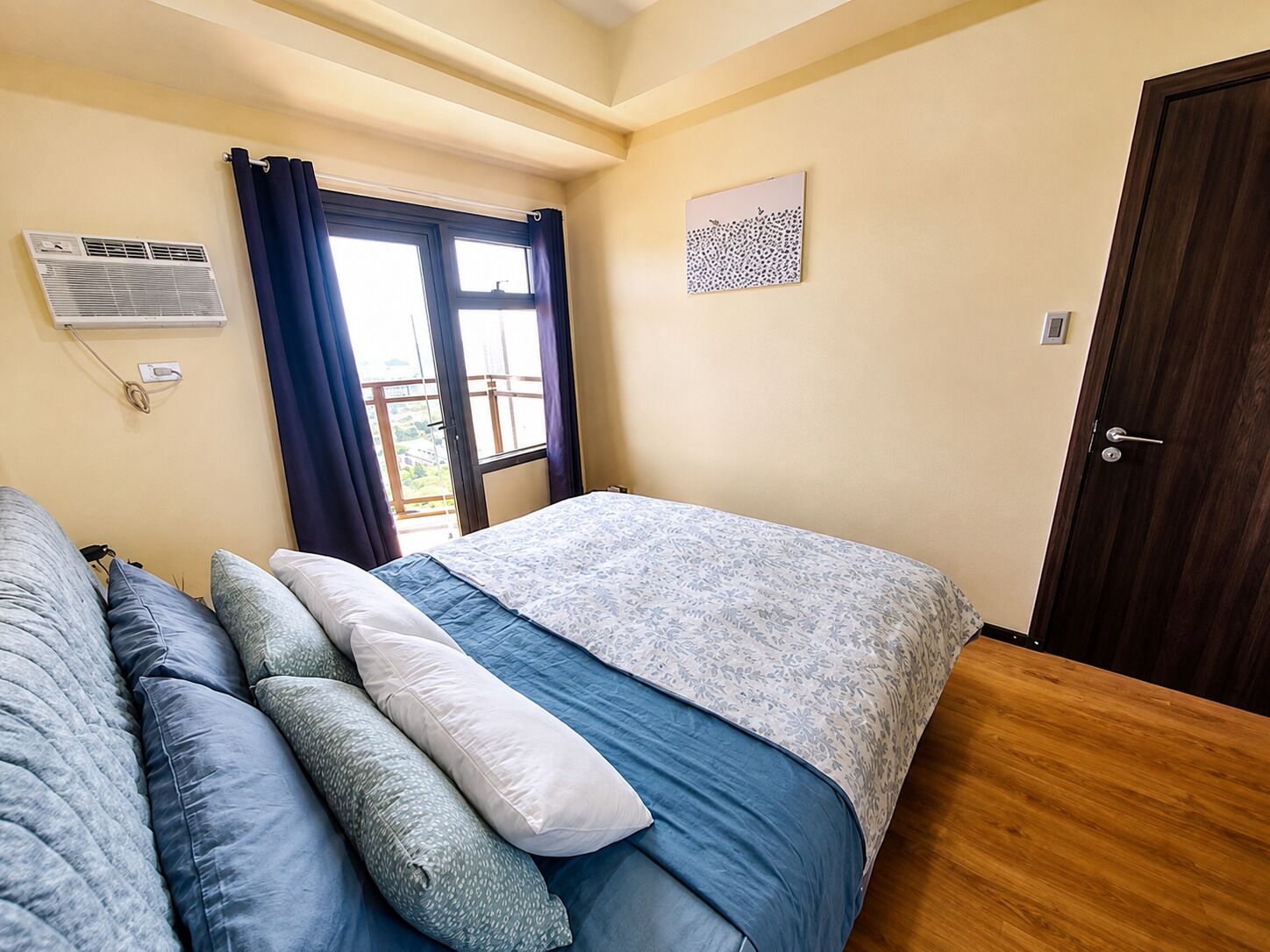 Ein Schlafzimmer mit Bett, einem Fenster mit Vorhängen, einer Tür und einer Wand-Klimaanlage.