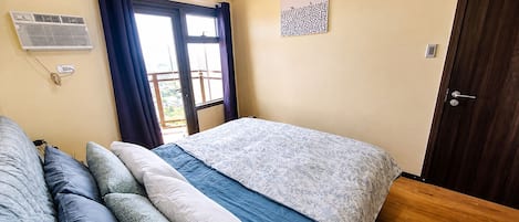 A bedroom with a bed, a window with curtains, a door, and a wall-mounted air conditioner.