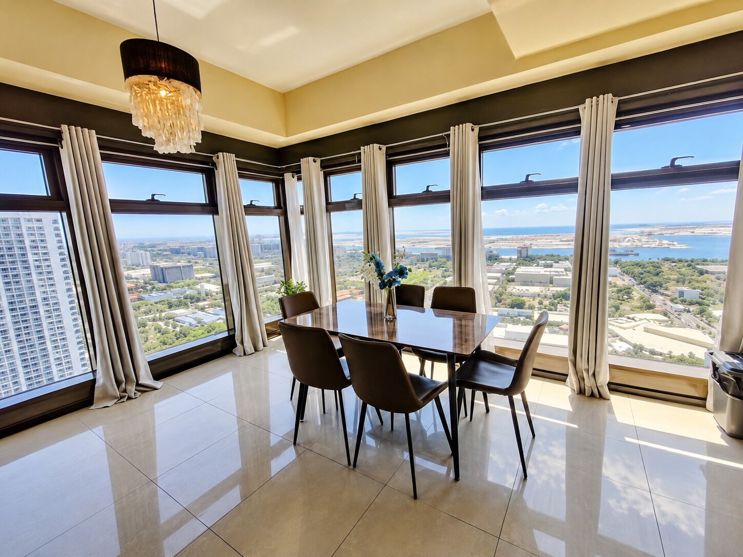 A modern dining area with a large table and chairs, large windows with curtains, and a city view.