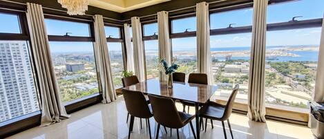 A modern dining area with a large table and chairs, large windows with curtains, and a city view.
