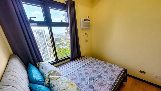 Ein Schlafzimmer mit einem Bett, einem fensterbelaubten Fenster, einer Wand-Klimaanlage und Blick auf eine Stadtlandschaft.