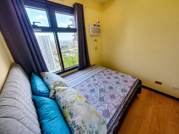 A bedroom with a bed, a window with curtains, a wall-mounted air conditioner, and a view of a cityscape.