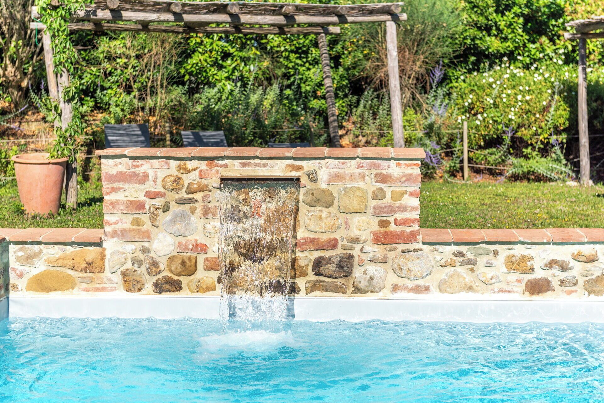 Un muro in pietra con una fontana, un pergolato in legno e un giardino con diverse piante.