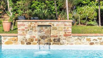 Un muro in pietra con una fontana, un pergolato in legno e un giardino con diverse piante.